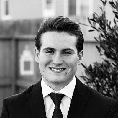 This is an alternate headshot for UW Tacoma Senior, Austin Donovan. Here Austin is wearing a suit in a black-and-white portrait framed by fencing and a tree.